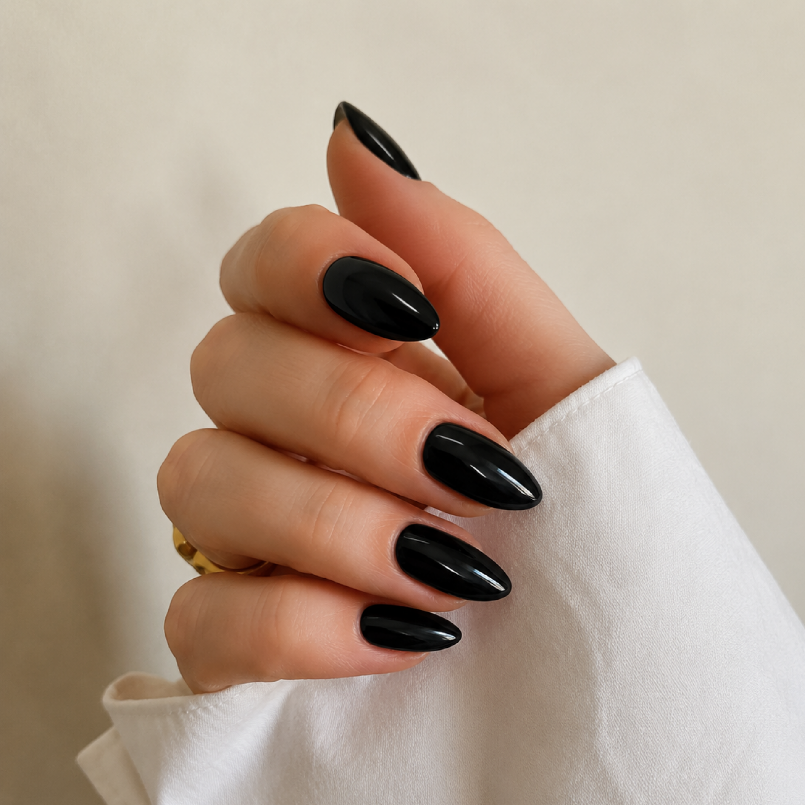 A close-up of a hand with glossy black almond-shaped nails, wearing a white long-sleeve shirt and a gold ring, against a soft beige background.