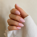 A close-up of a hand with a glossy, soft pink manicure, wearing a white textured sleeve and a small gold ring, set against a neutral beige background.