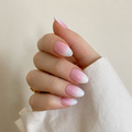 A close-up of a hand with long almond-shaped nails styled in a soft pink and white ombre gradient, wearing a white long-sleeve shirt against a warm beige background.