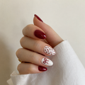 A close-up of a hand with manicured nails, featuring a mix of deep burgundy and white nail art designs including polka dots and a bow, wearing a white sleeve and a small gold ring.