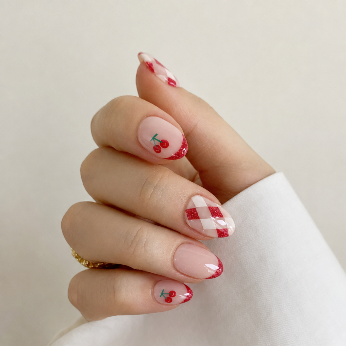 A close-up of a hand with manicured nails featuring a cherry and red-and-white gingham pattern, wearing a white long-sleeve top against a soft beige background.