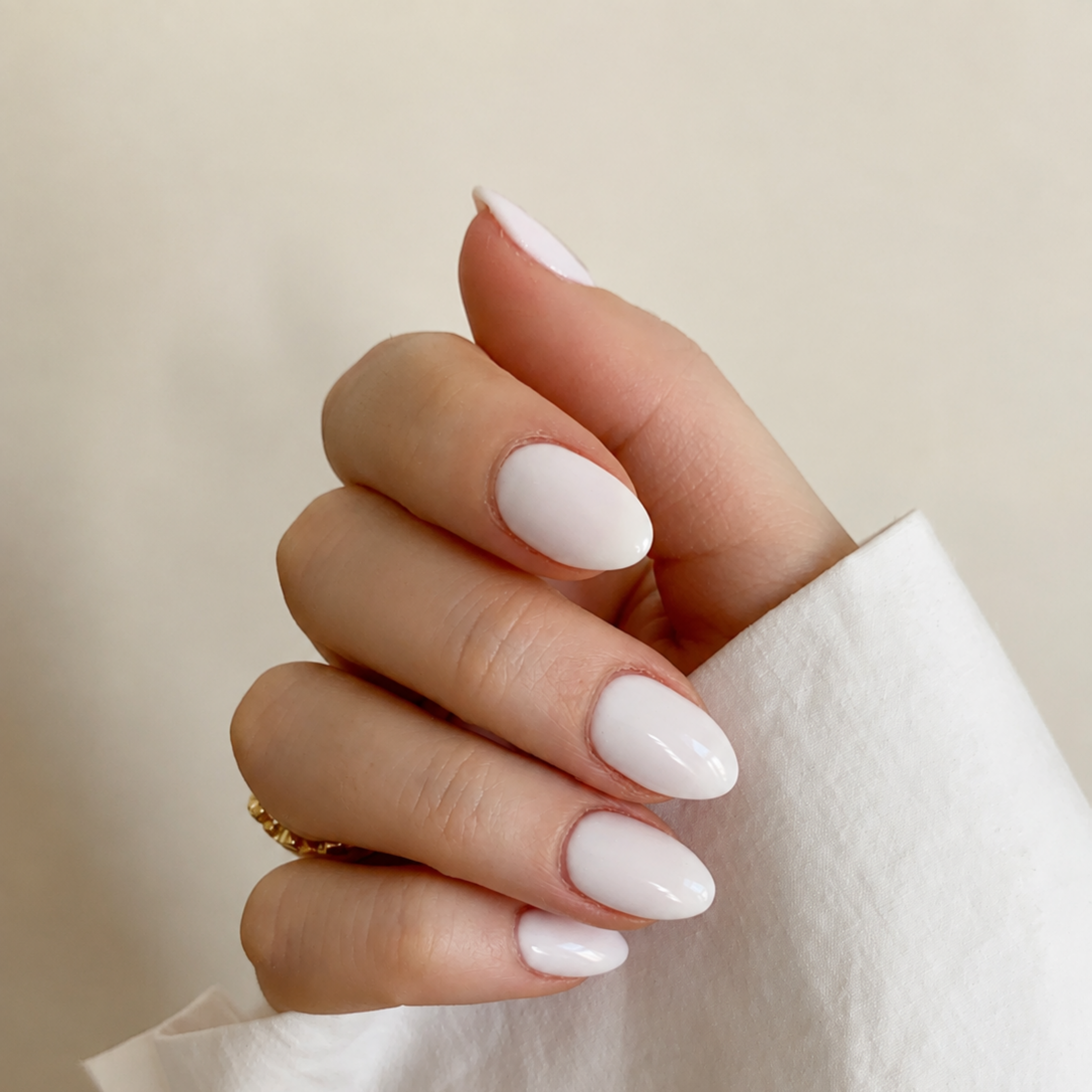 A close-up of a hand with a glossy, solid pale pink nail polish, wearing a white sleeve and a small gold ring against a soft beige background.