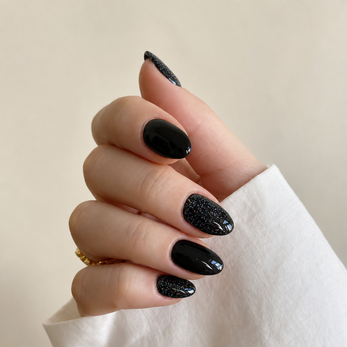 A close-up of a hand with manicured nails painted in glossy black and black glitter, wearing a white sleeve and a small gold ring against a soft beige background.