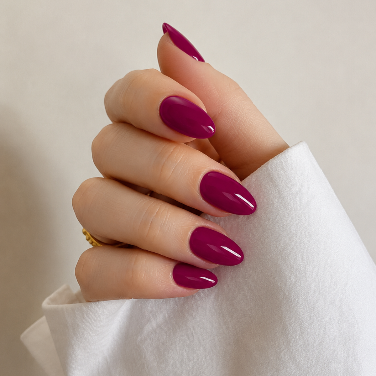 A close-up of a hand with glossy, almond-shaped nails painted in a deep berry purple shade, wearing a white sleeve and a gold ring against a soft beige background.
