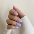 A close-up of a hand with a glossy lavender purple manicure, wearing a white sleeve and a small gold ring, against a soft beige background.