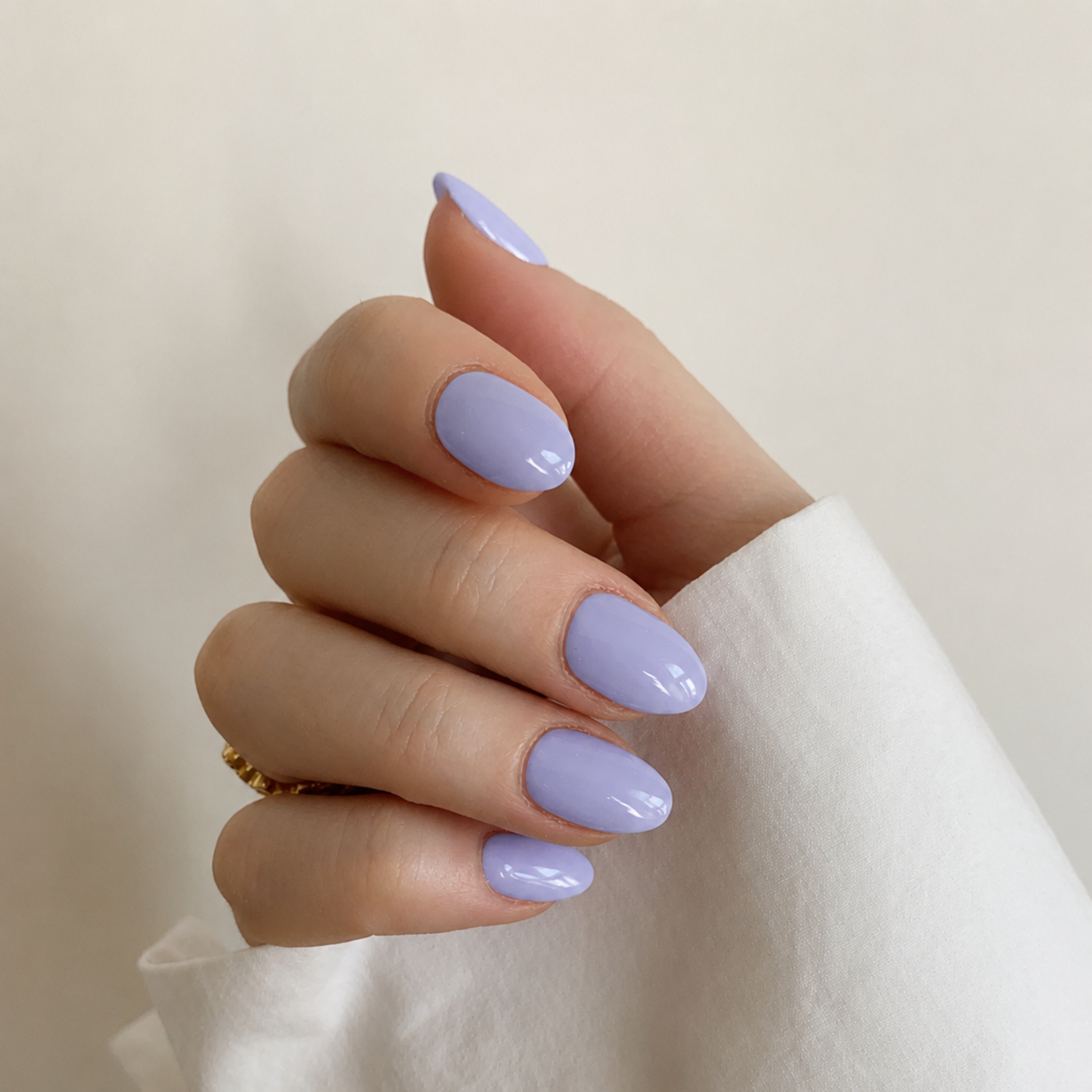 A close-up of a hand with a glossy lavender purple manicure, wearing a white sleeve and a small gold ring, against a soft beige background.