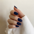 A close-up of a hand with neatly manicured, almond-shaped nails painted in a glossy deep navy blue, resting against a soft beige background with a white sleeve visible.