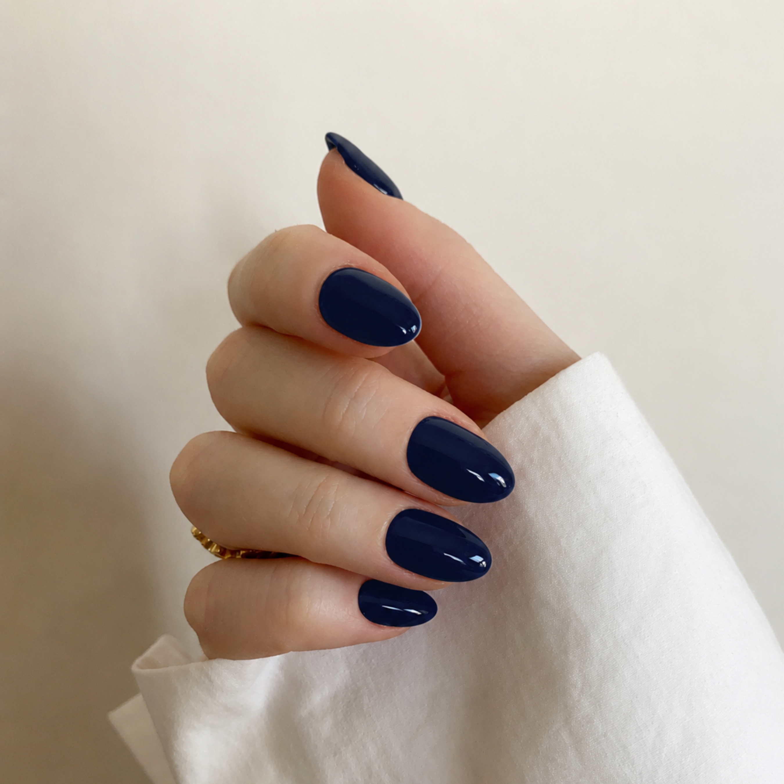 A close-up of a hand with neatly manicured, almond-shaped nails painted in a glossy deep navy blue, resting against a soft beige background with a white sleeve visible.