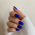A close-up of a hand with glossy, almond-shaped nails painted in a vibrant royal blue color, wearing a white sleeve and a small gold ring against a soft beige background.