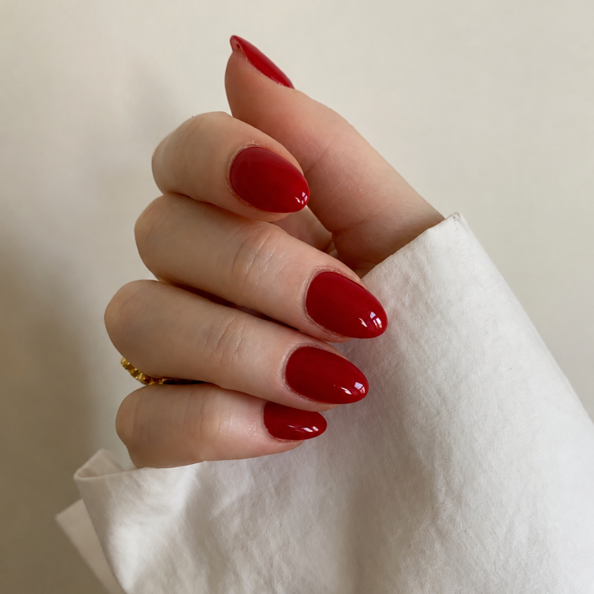 A close-up of a hand with glossy, almond-shaped nails painted in a vibrant deep red, resting against a white sleeve and a neutral beige background.