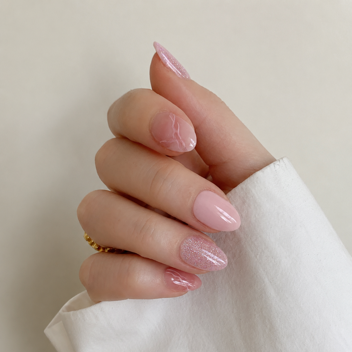 A close-up of a hand with manicured nails in a soft pink and glittery pink ombre style, wearing a white sleeve and a small gold ring against a plain beige background.