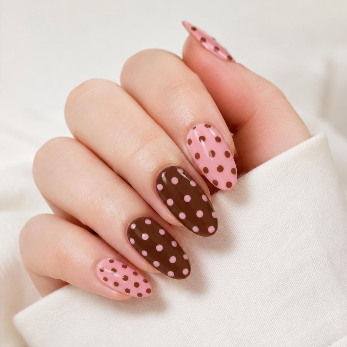 A close-up of a hand with almond-shaped nails styled in a pink and brown polka dot pattern, wearing a white long-sleeve top against a soft white background.